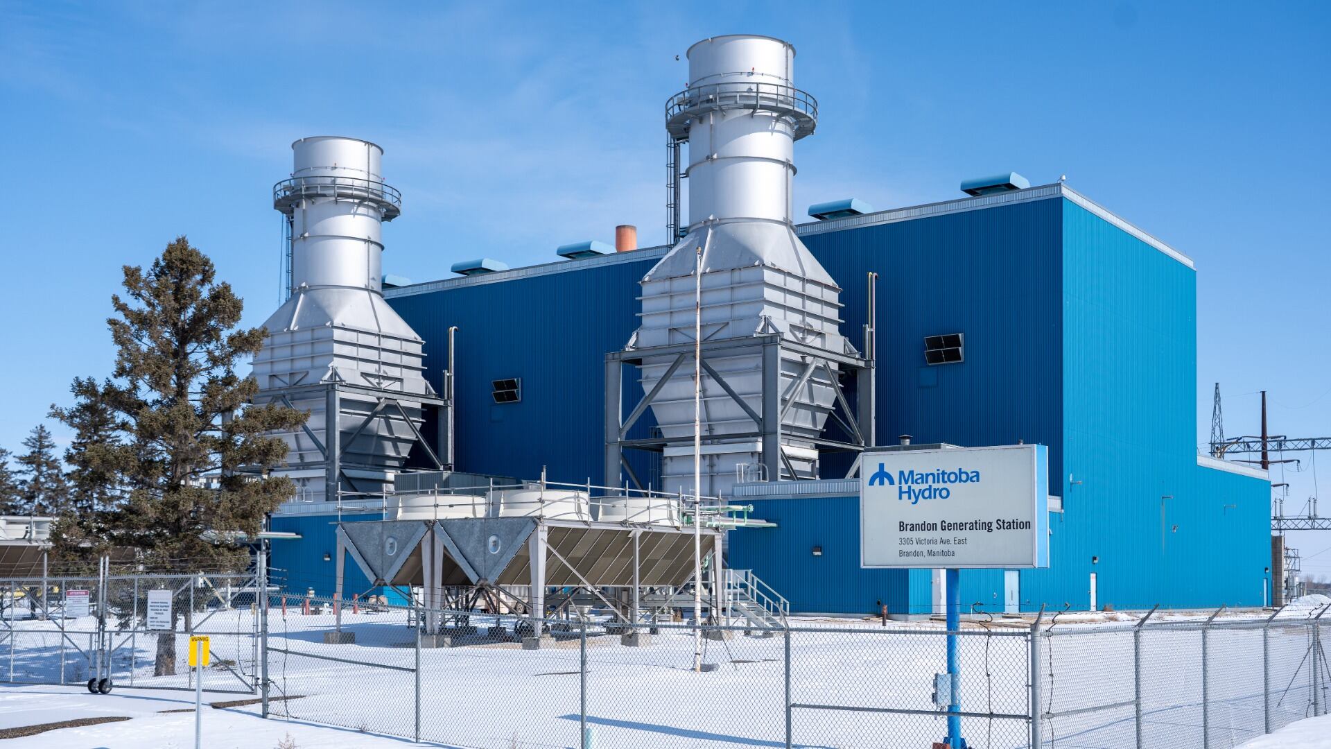 The Brandon Generating Station: a blue industrial building with two silver gas turbine exhaust stacks behind a chain-link fence in snow, with a Manitoba Hydro sign in front