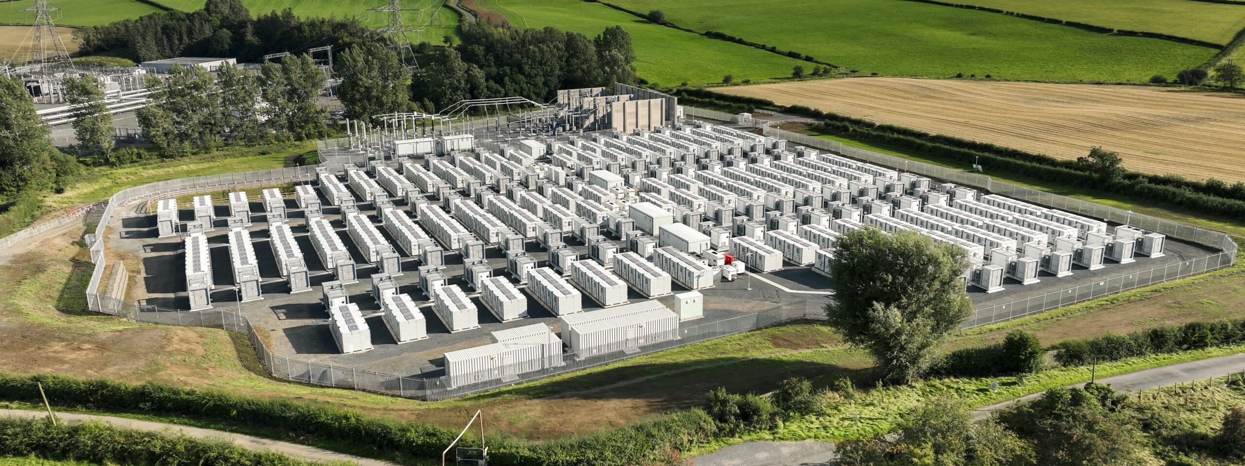Aerial view of white battery container rows in a fenced compound beside transmission pylons and farmland, Kilmarnock South BESS, Scotland.
