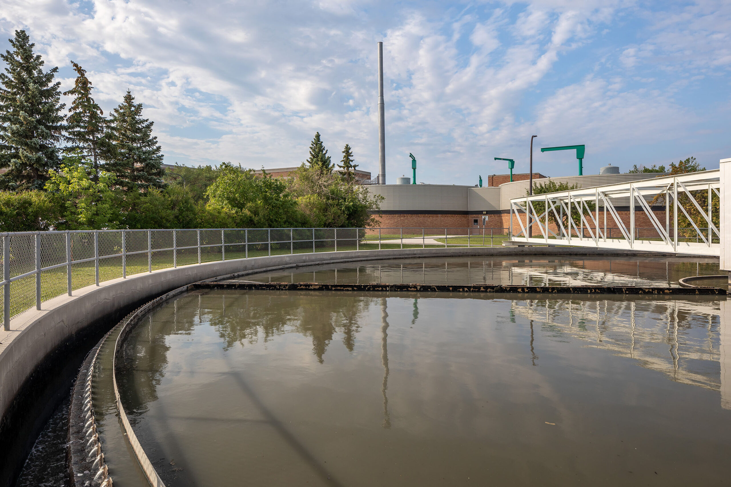 A circular clarifier pool of warm treated water at the North End Water Pollution Control Centre, with the plant buildings and stack behind